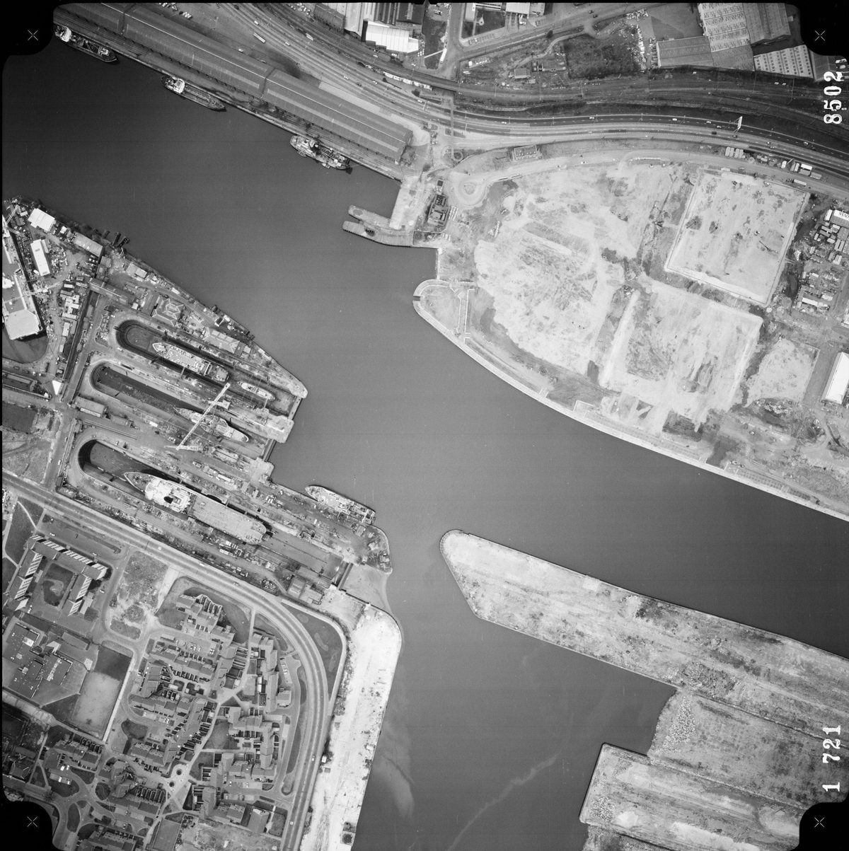 A large-scale black and white aerial photo of Glasgow. The river from the previous images now occupies almost the whole frame. At the left side, ships are clearly visible docked next to the river. There are blocks of flats in the bottom left corner.