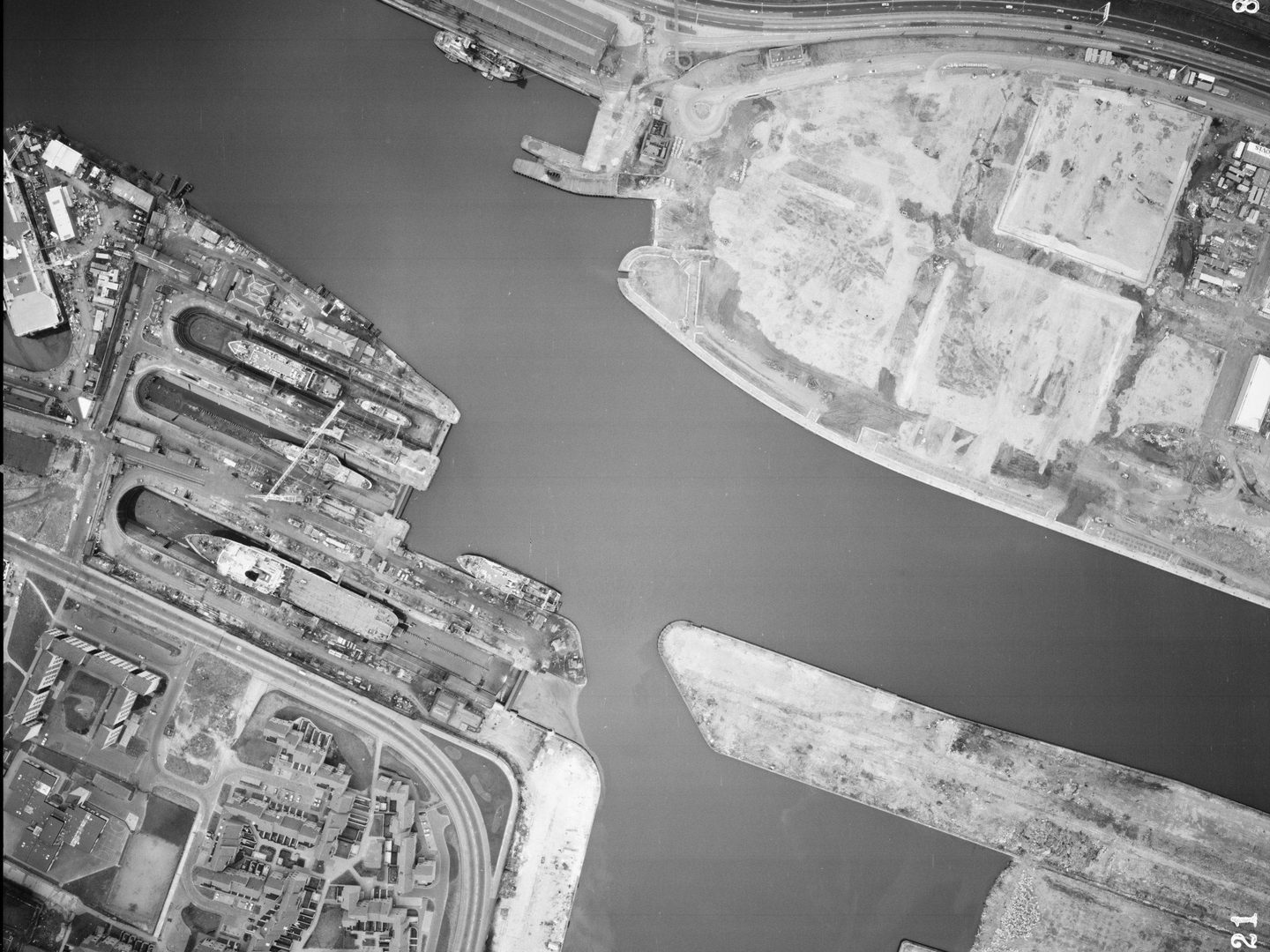 A large-scale black and white aerial photo of Glasgow. The river from the previous images now occupies almost the whole frame. At the left side, ships are clearly visible docked next to the river. There are blocks of flats in the bottom left corner.