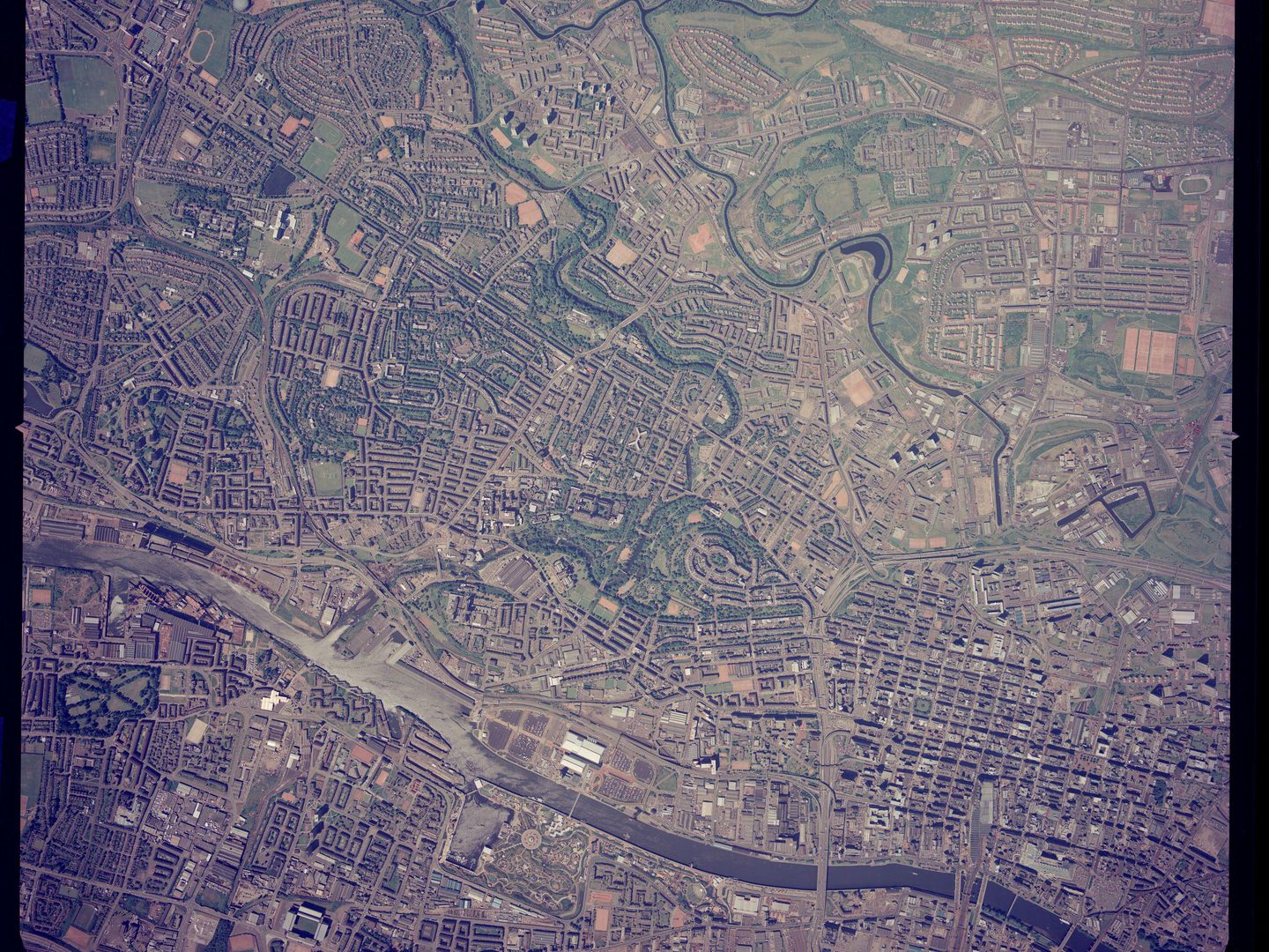 A small-scale colour aerial photo of Glasgow. Grids of streets are visible across the image, interspersed with green parks to the left, and with extensive green spaces in the top right corner. A river runs along the bottom edge.