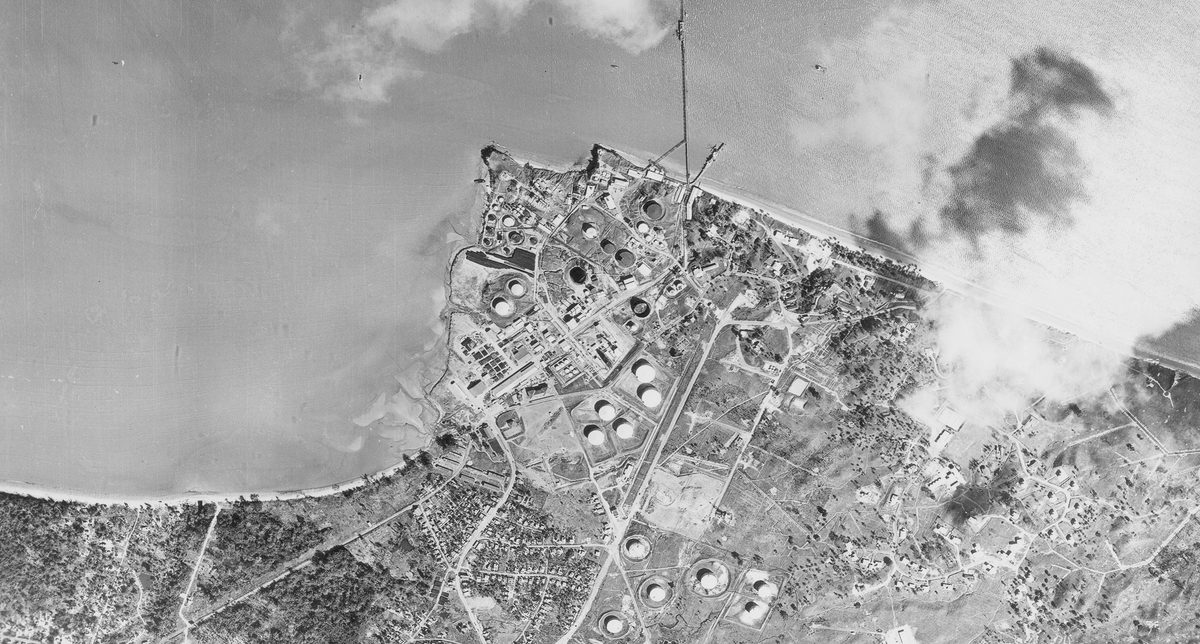 A historic black and white vertical aerial photo of an industrial area with large oil tanks on a shoreline.