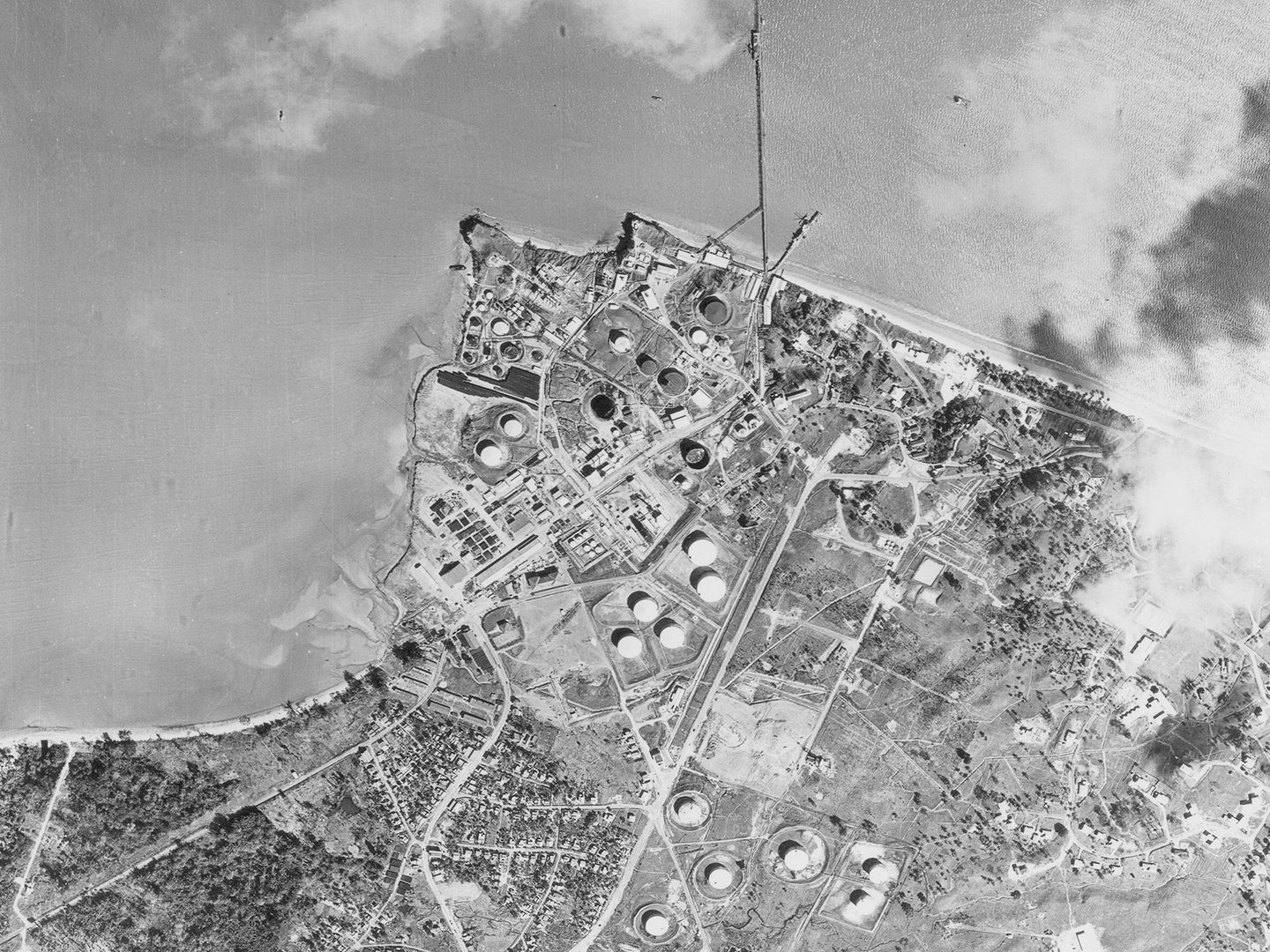 A historic black and white vertical aerial photo of an industrial area with large oil tanks on a shoreline.