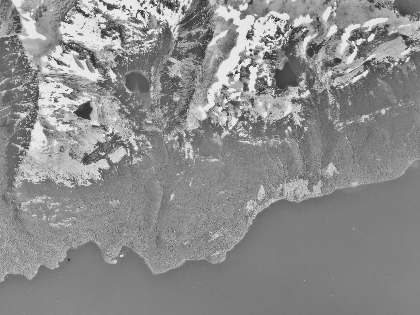 A historic black and white vertical aerial photo of a ragged shoreline. A lake stretches along the bottom of the image, with the land rising to snowy peaks along the top.