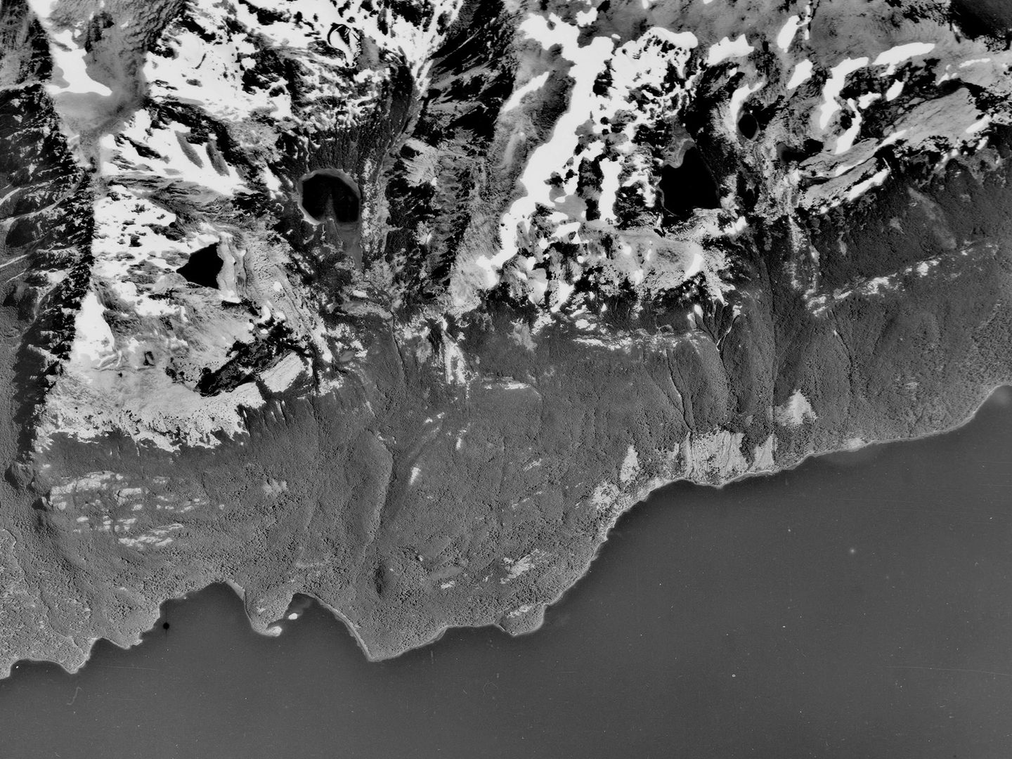 A historic black and white vertical aerial photo of a ragged shoreline. A lake stretches along the bottom of the image, with the land rising to snowy peaks along the top.