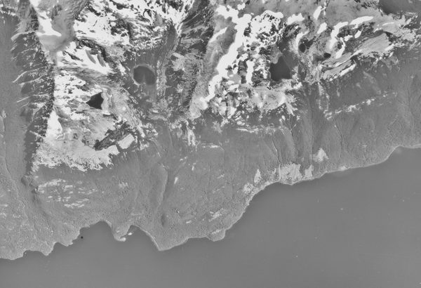 A historic black and white vertical aerial photo of a ragged shoreline. A lake stretches along the bottom of the image, with the land rising to snowy peaks along the top.
