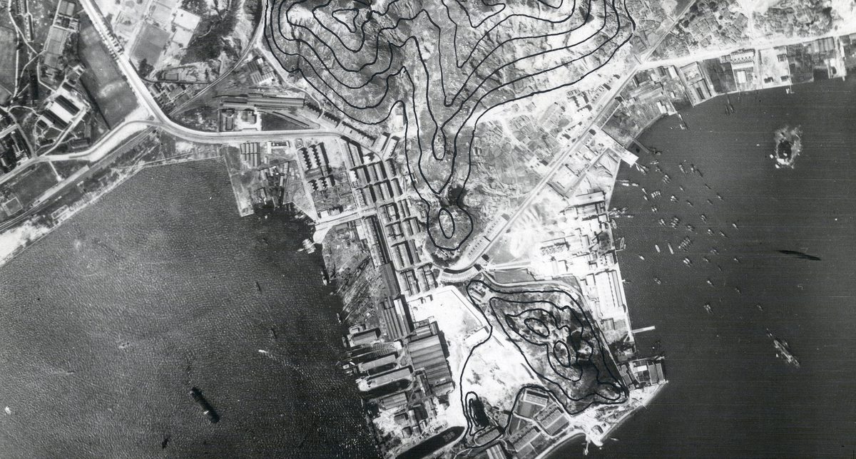 A historic black and white vertical aerial photo of Hong Kong, with densely packed buildings covering a promontory surrounded by sea. Several vessels are on the water. The photo has been marked with black lines to show contours.