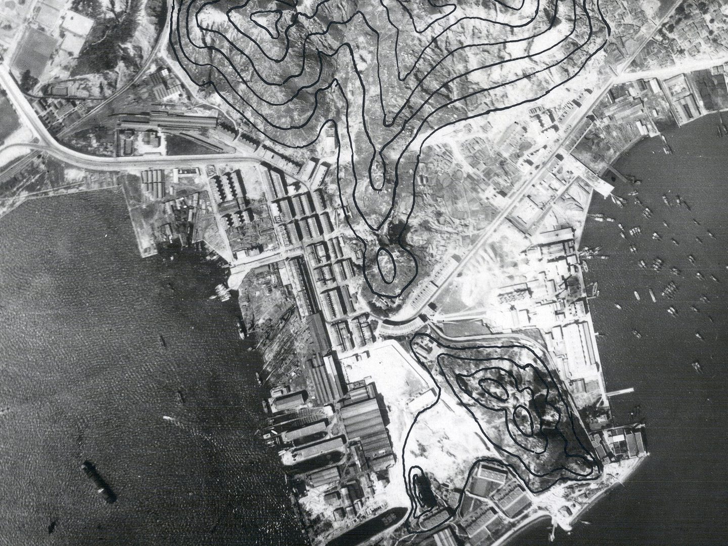 A historic black and white vertical aerial photo of Hong Kong, with densely packed buildings covering a promontory surrounded by sea. Several vessels are on the water. The photo has been marked with black lines to show contours.