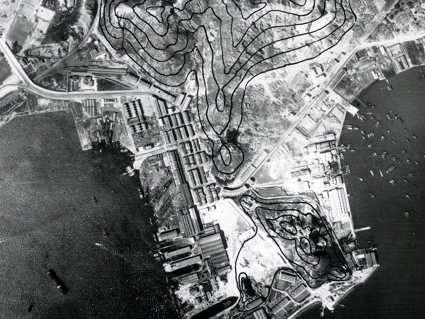 A historic black and white vertical aerial photo of Hong Kong, with densely packed buildings covering a promontory surrounded by sea. Several vessels are on the water. The photo has been marked with black lines to show contours.