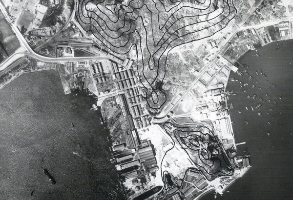 A historic black and white vertical aerial photo of Hong Kong, with densely packed buildings covering a promontory surrounded by sea. Several vessels are on the water. The photo has been marked with black lines to show contours.