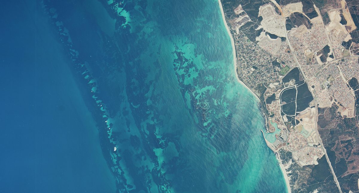 A historic colour vertical aerial photo of a coastline. The left two-thirds of the image are filled with a very blue sea. The shore to the right looks dry and arid.