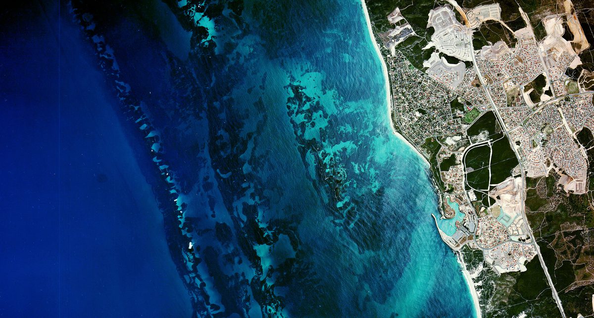 A historic colour vertical aerial photo of a coastline. The left two-thirds of the image are filled with a very blue sea. The shore to the right looks dry and arid.