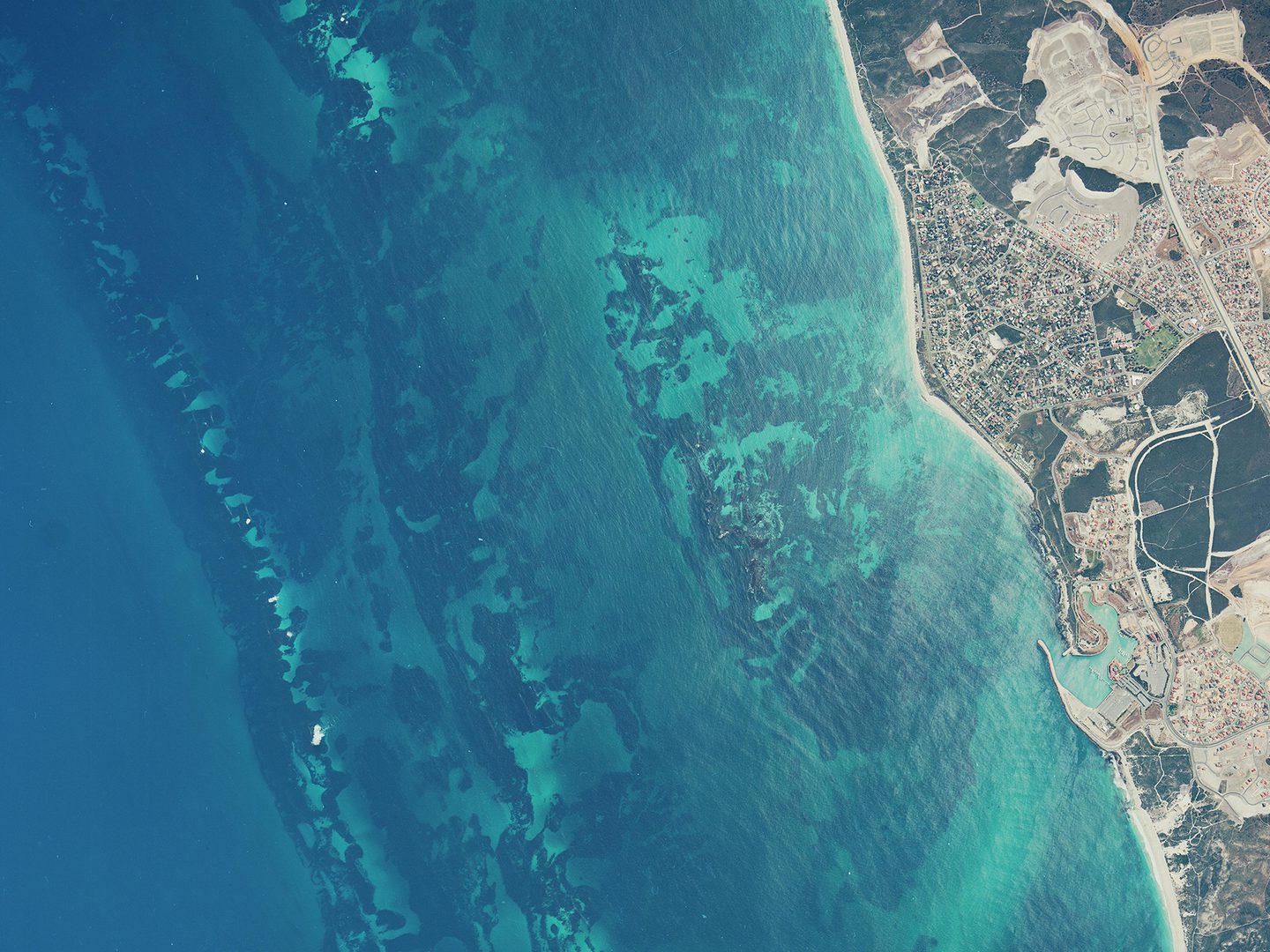 A historic colour vertical aerial photo of a coastline. The left two-thirds of the image are filled with a very blue sea. The shore to the right looks dry and arid.