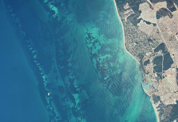 A historic colour vertical aerial photo of a coastline. The left two-thirds of the image are filled with a very blue sea. The shore to the right looks dry and arid.