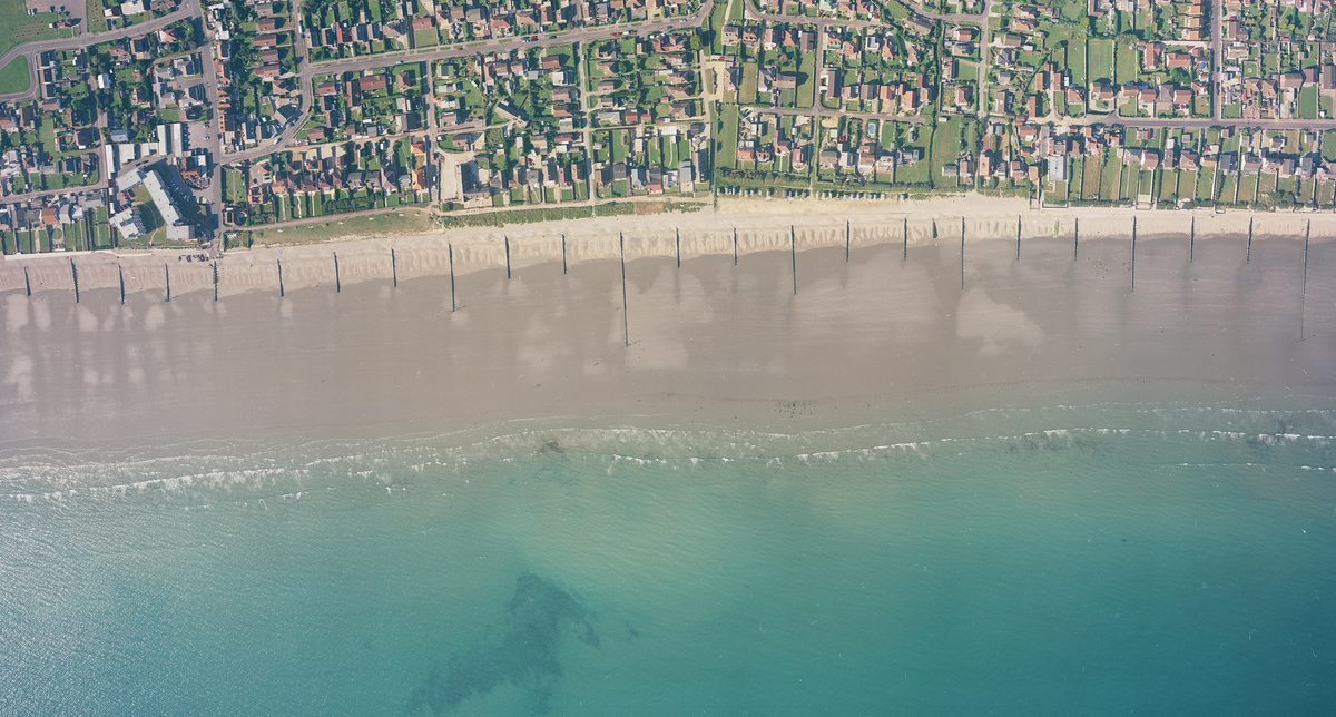 A historic colour vertical aerial photo of a shoreline. Along the bottom of the image is a blue sea. A wide beach interspersed with groynes runs along the sea, with the houses of a town inland from the beach.