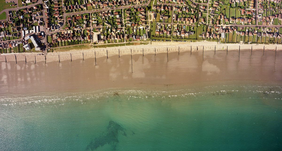 A historic colour vertical aerial photo of a shoreline. Along the bottom of the image is a blue sea. A wide beach interspersed with groynes runs along the sea, with the houses of a town inland from the beach.