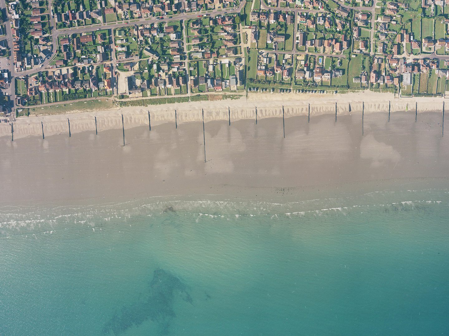 A historic colour vertical aerial photo of a shoreline. Along the bottom of the image is a blue sea. A wide beach interspersed with groynes runs along the sea, with the houses of a town inland from the beach.