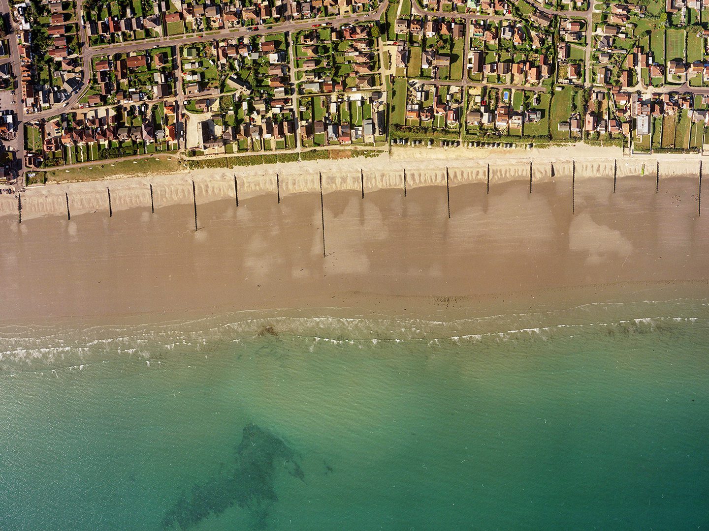 A historic colour vertical aerial photo of a shoreline. Along the bottom of the image is a blue sea. A wide beach interspersed with groynes runs along the sea, with the houses of a town inland from the beach.