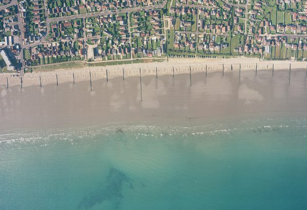 A historic colour vertical aerial photo of a shoreline. Along the bottom of the image is a blue sea. A wide beach interspersed with groynes runs along the sea, with the houses of a town inland from the beach.