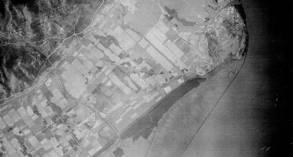 A historic black and white vertical aerial photo of agricultural land along a coastline. The sea is to the right of the image, and some hills are visible in the top left corner.