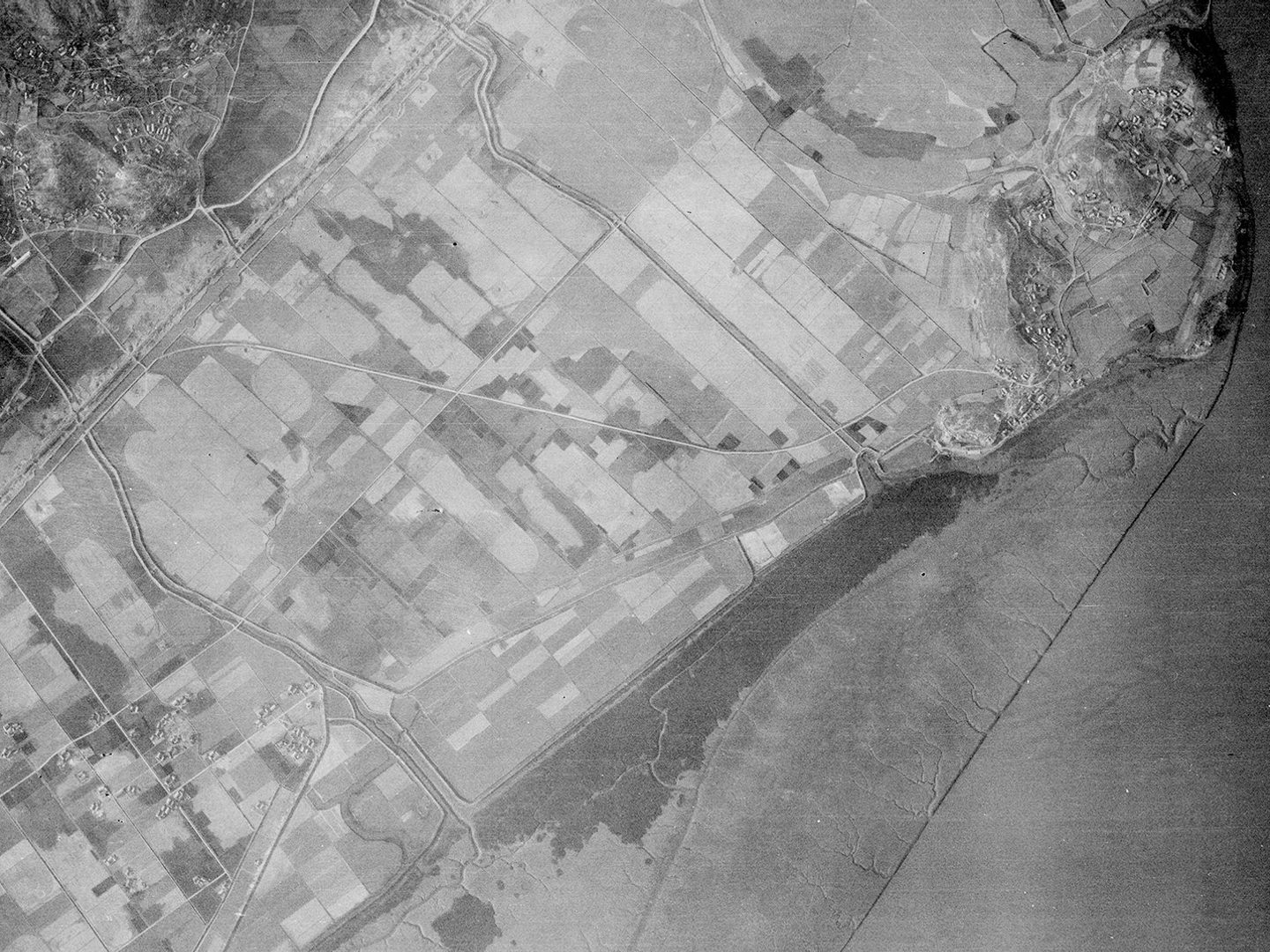 A historic black and white vertical aerial photo of agricultural land along a coastline. The sea is to the right of the image, and some hills are visible in the top left corner.