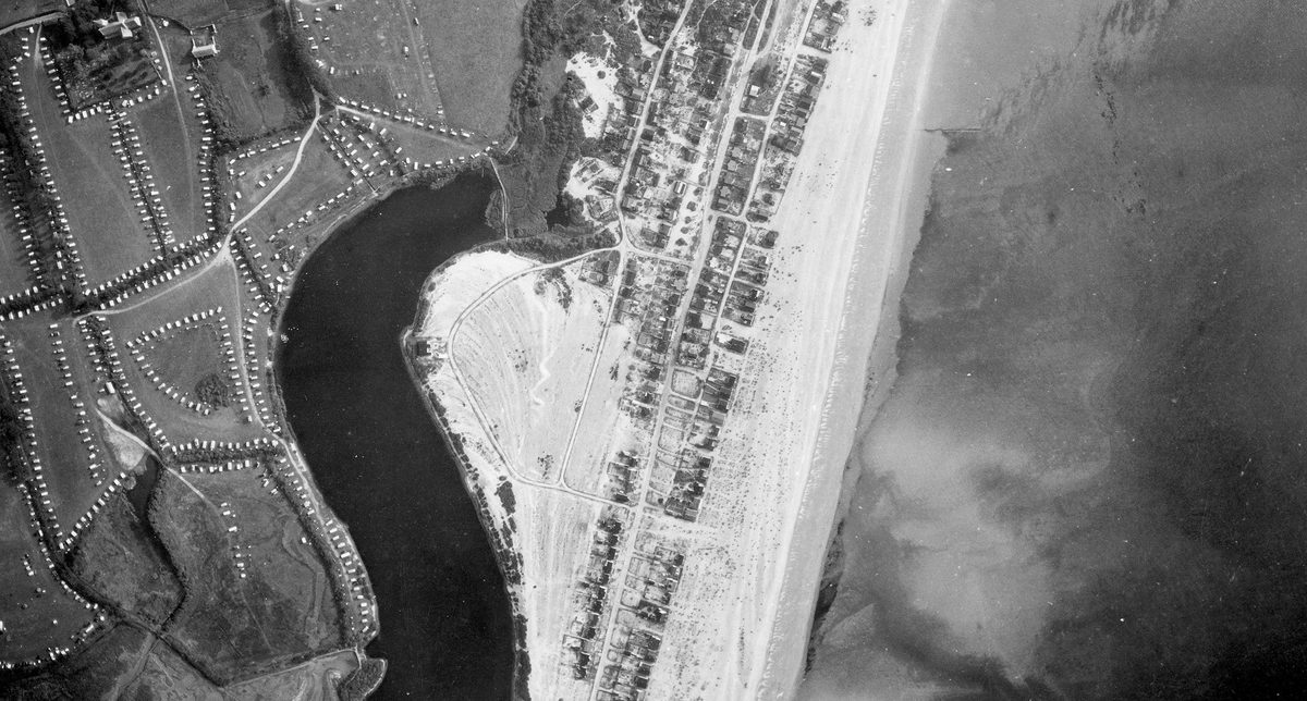 A historic black and white vertical aerial photo of a coastline. To the right is the sea, separated from a small town by a beach. Inland is a caravan park and a dark body of water.