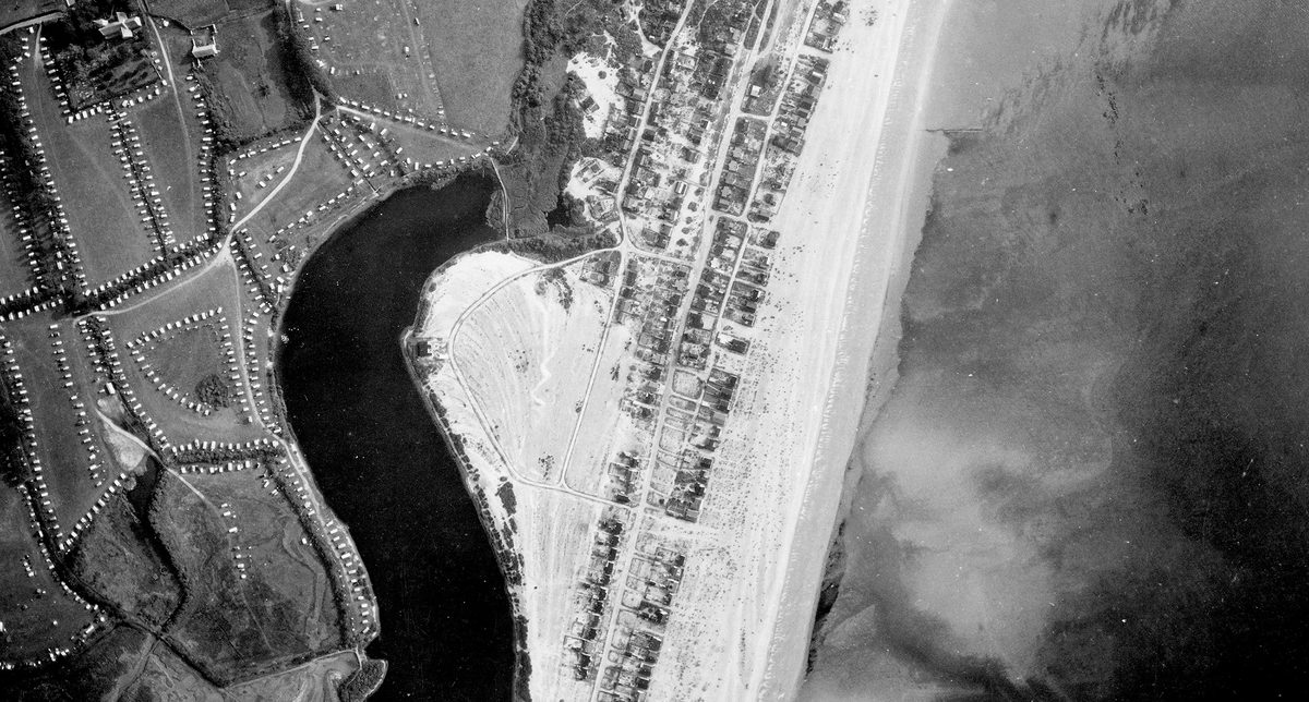A historic black and white vertical aerial photo of a coastline. To the right is the sea, separated from a small town by a beach. Inland is a caravan park and a dark body of water.
