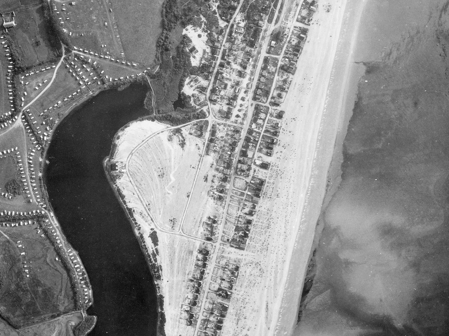 A historic black and white vertical aerial photo of a coastline. To the right is the sea, separated from a small town by a beach. Inland is a caravan park and a dark body of water.