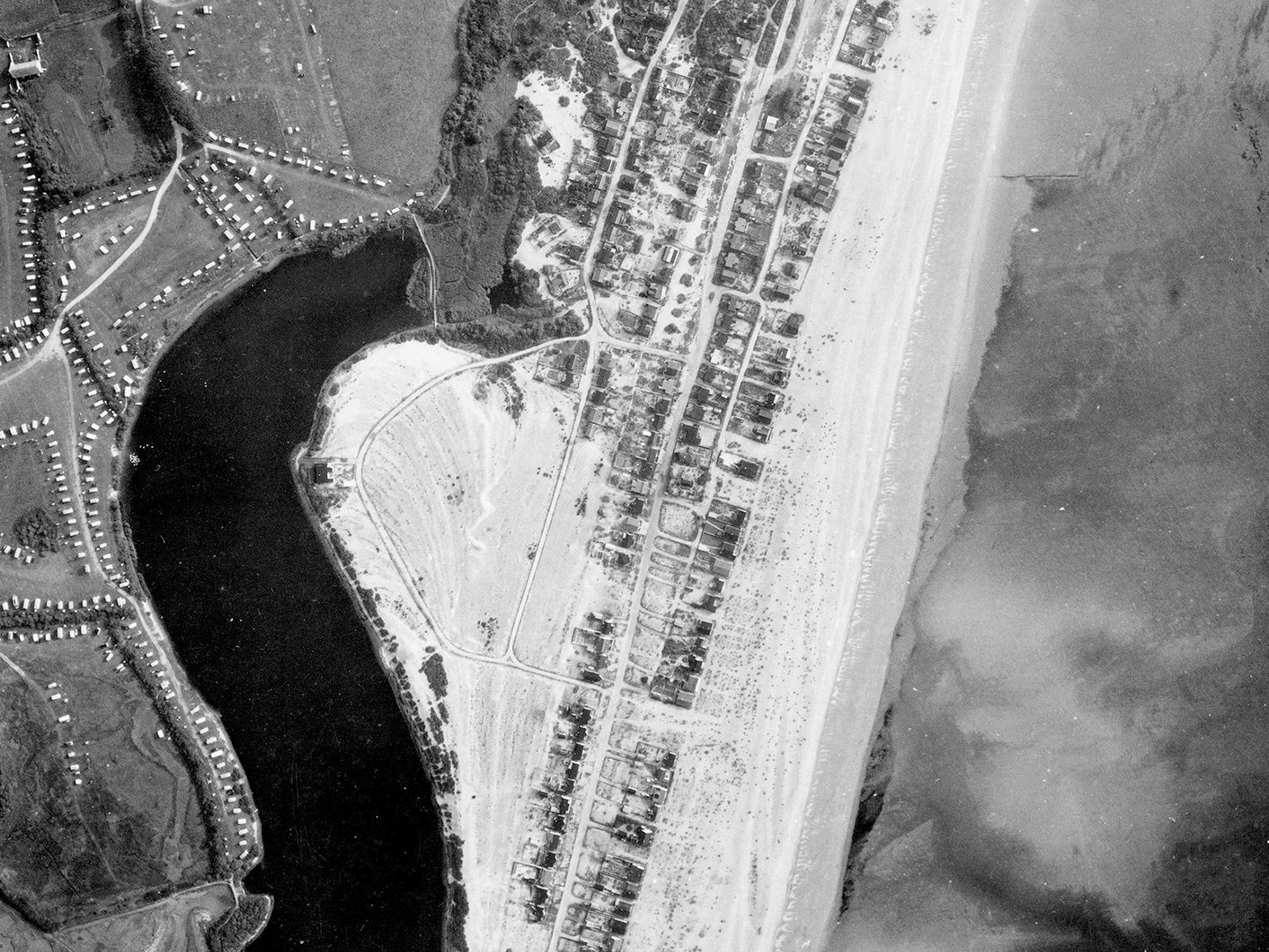 A historic black and white vertical aerial photo of a coastline. To the right is the sea, separated from a small town by a beach. Inland is a caravan park and a dark body of water.