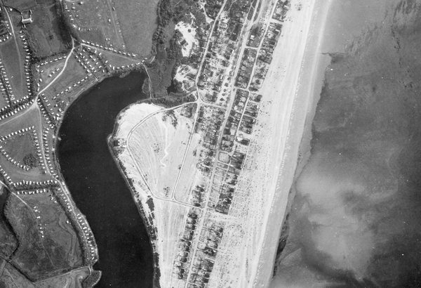 A historic black and white vertical aerial photo of a coastline. To the right is the sea, separated from a small town by a beach. Inland is a caravan park and a dark body of water.
