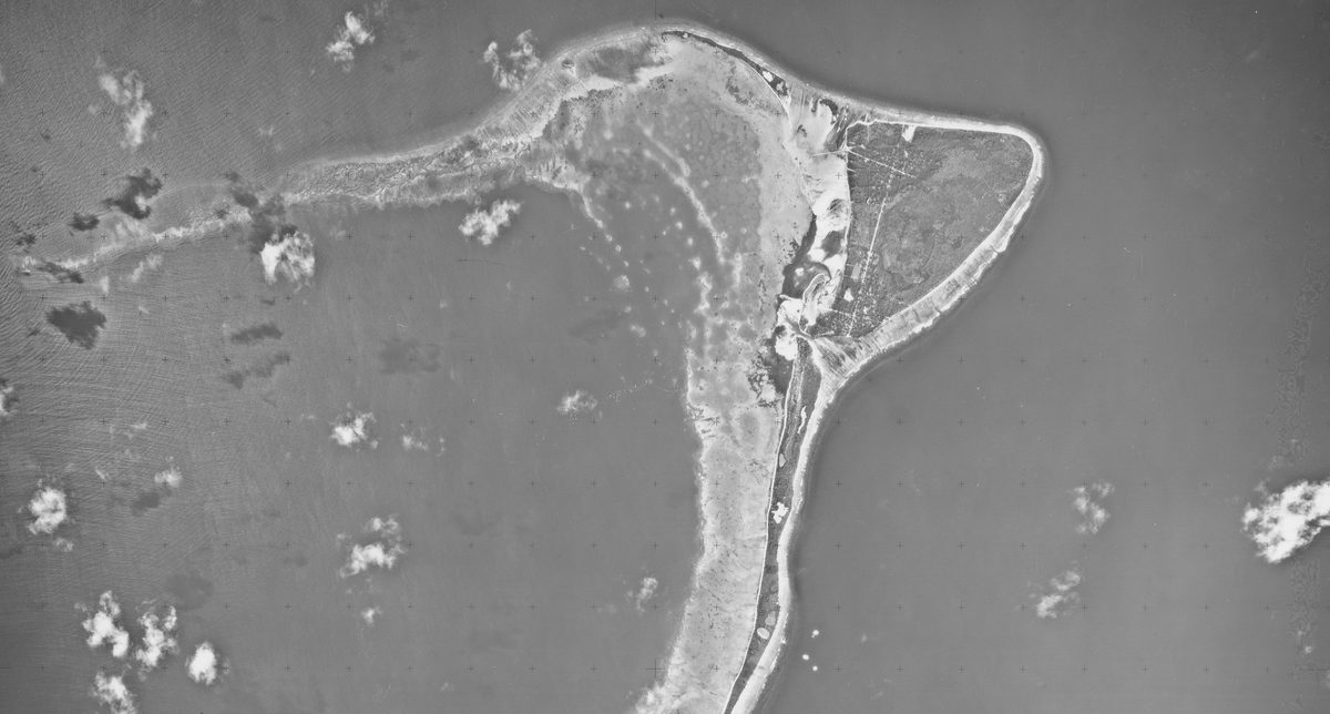 A historic black and white vertical aerial photo of a crescent-shaped island, fringed by beaches and surrounded by the sea. A few small white clouds are visible.