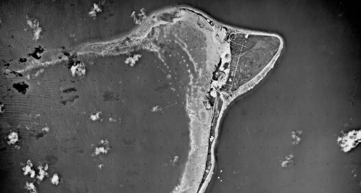 A historic black and white vertical aerial photo of a crescent-shaped island, fringed by beaches and surrounded by the sea. A few small white clouds are visible.
