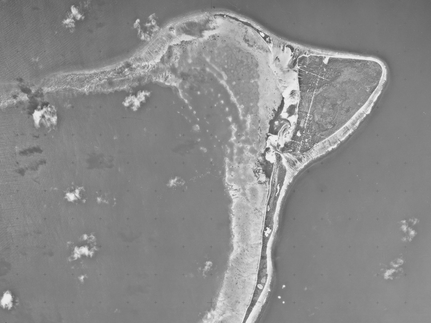 A historic black and white vertical aerial photo of a crescent-shaped island, fringed by beaches and surrounded by the sea. A few small white clouds are visible.