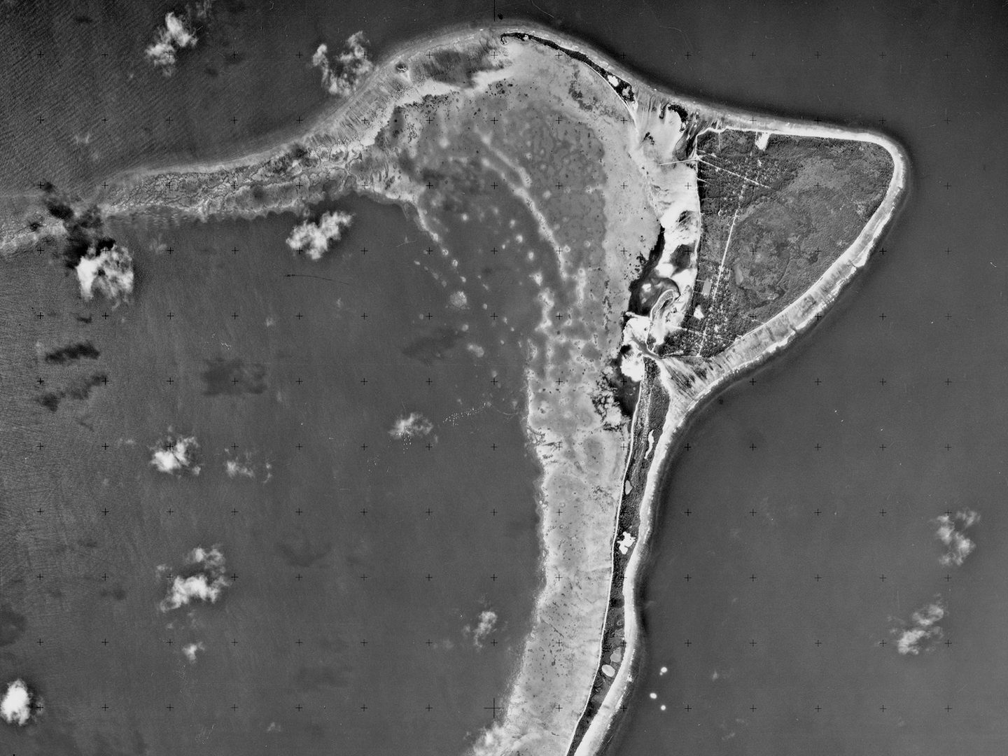 A historic black and white vertical aerial photo of a crescent-shaped island, fringed by beaches and surrounded by the sea. A few small white clouds are visible.