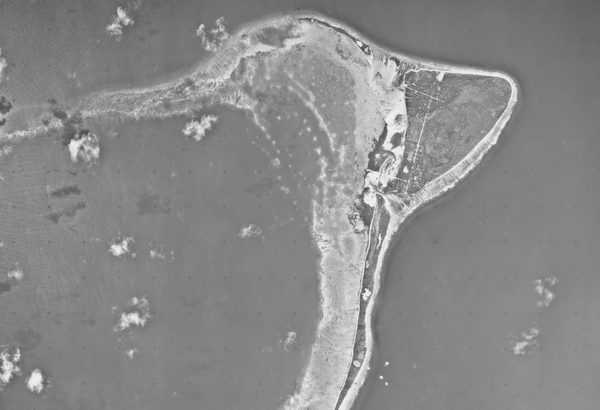 A historic black and white vertical aerial photo of a crescent-shaped island, fringed by beaches and surrounded by the sea. A few small white clouds are visible.