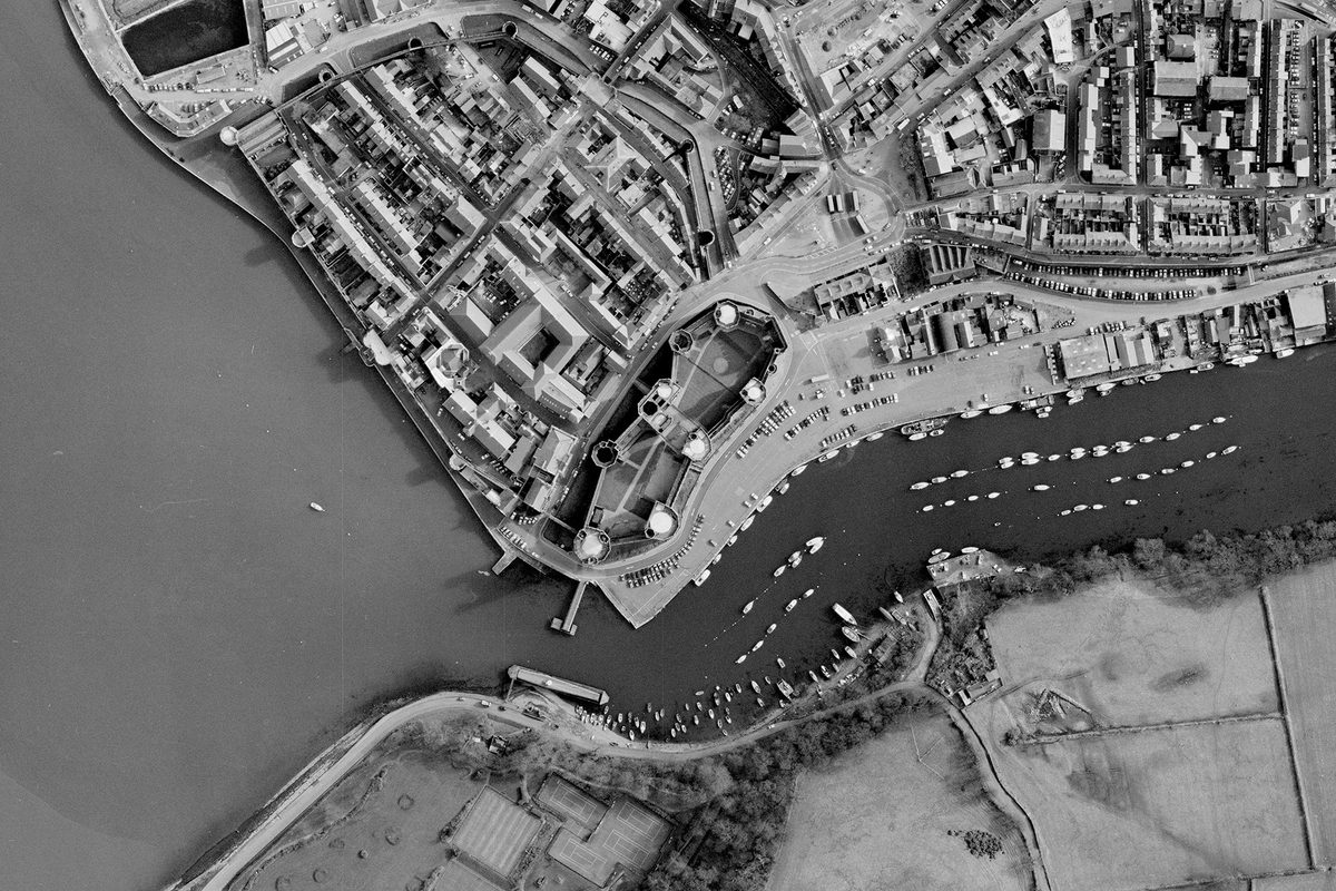 A historic black and white vertical aerial photo of Caernarfon Castle with 7 large towers in its curtain wall and a large gatehouse. To the left of the castle is a walled town. The town borders the sea. Along the bottom of the image is a river on which are lots of small boats.