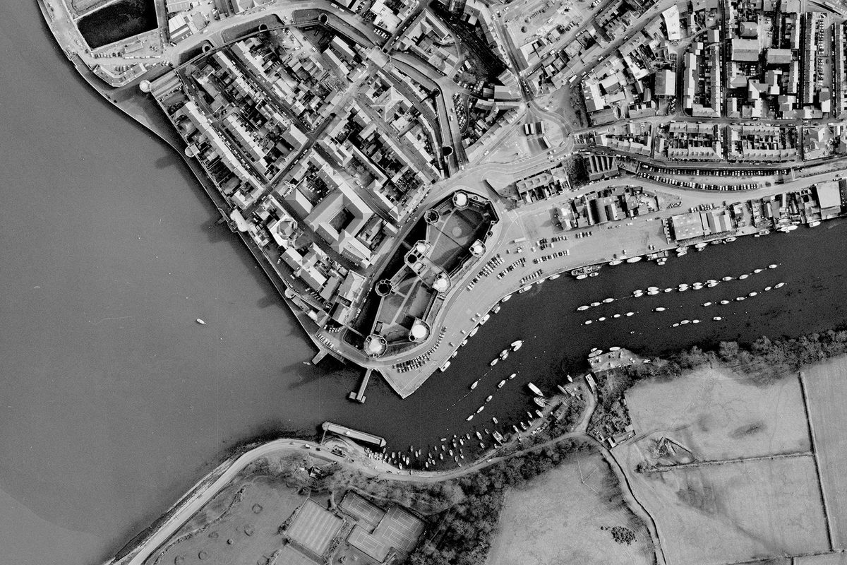 A historic black and white vertical aerial photo of Caernarfon Castle with 7 large towers in its curtain wall and a large gatehouse. To the left of the castle is a walled town. The town borders the sea. Along the bottom of the image is a river on which are lots of small boats.