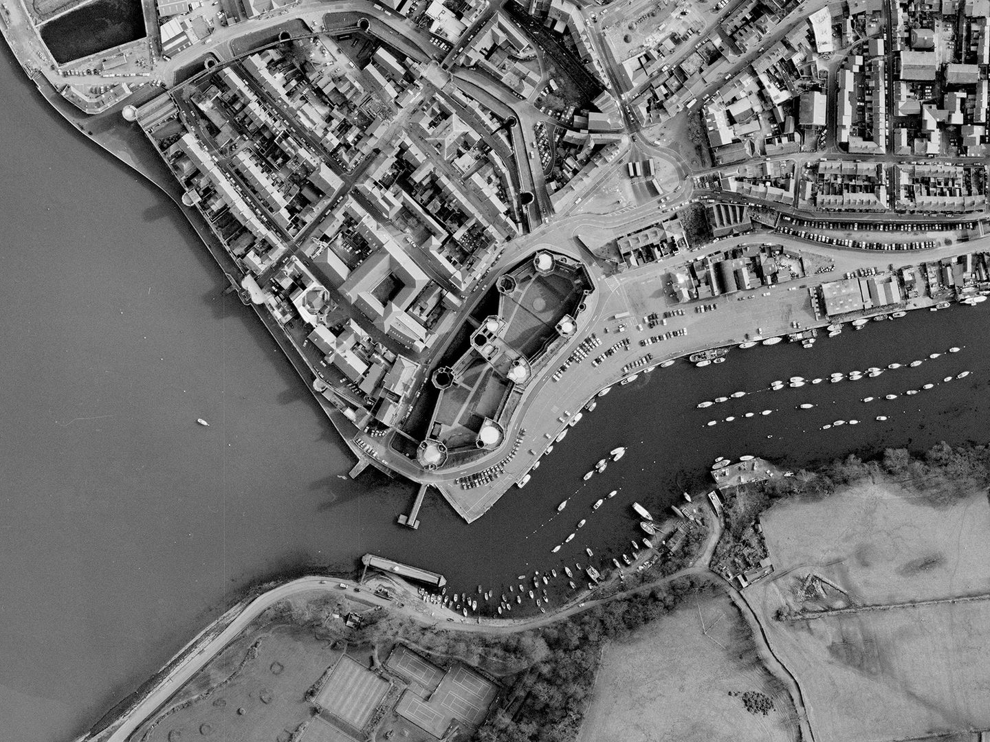 A historic black and white vertical aerial photo of Caernarfon Castle with 7 large towers in its curtain wall and a large gatehouse. To the left of the castle is a walled town. The town borders the sea. Along the bottom of the image is a river on which are lots of small boats.