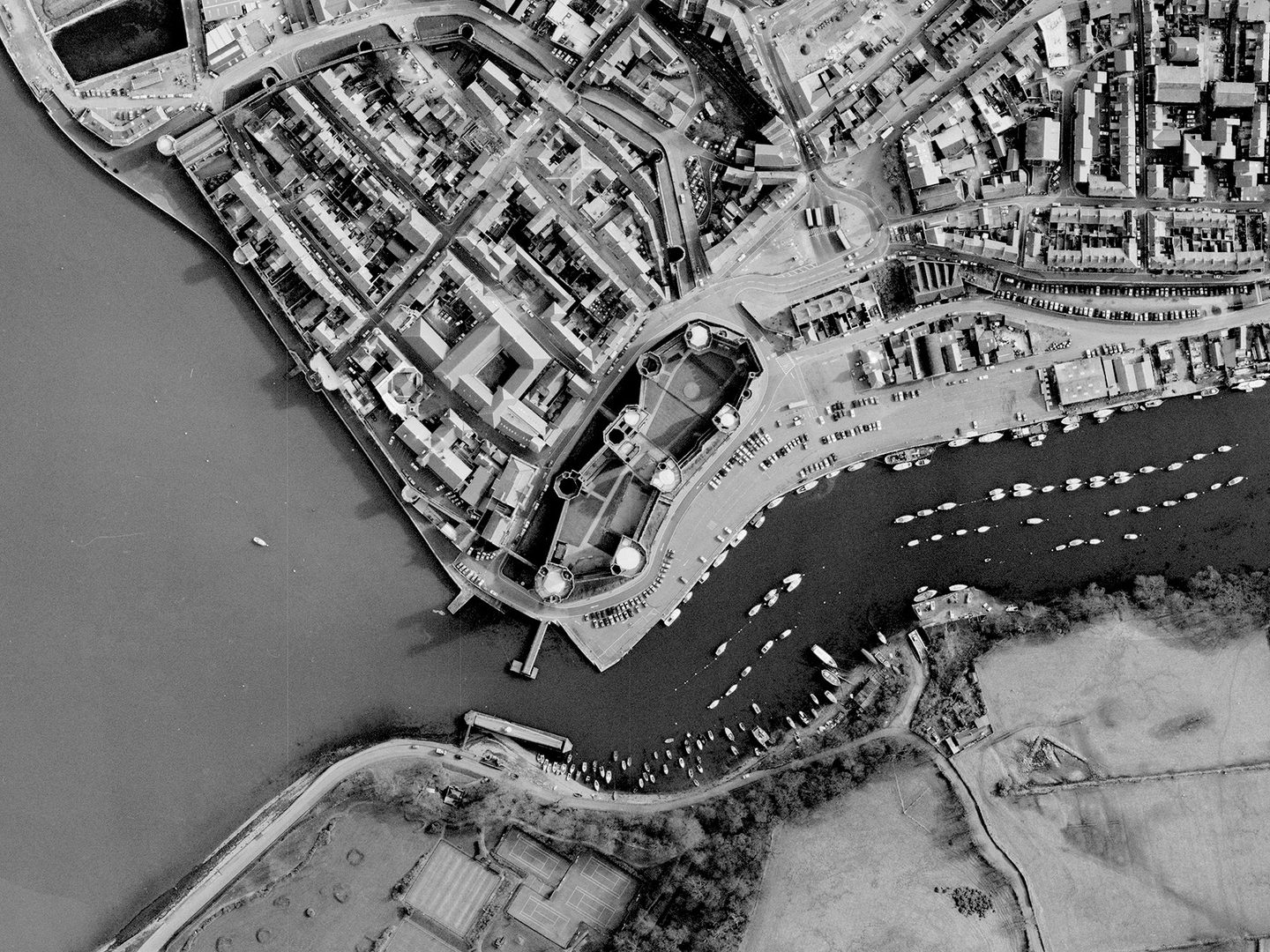 A historic black and white vertical aerial photo of Caernarfon Castle with 7 large towers in its curtain wall and a large gatehouse. To the left of the castle is a walled town. The town borders the sea. Along the bottom of the image is a river on which are lots of small boats.