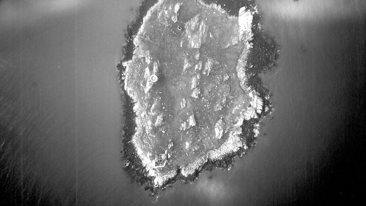 A historic black and white vertical aerial photo of a roughly oval-shaped island. A circular tower is visible to the left of centre, with a rectangular building complex at the bottom tip of the island.
