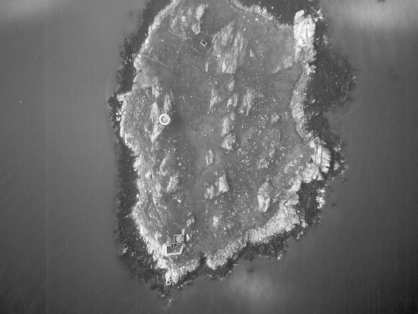 A historic black and white vertical aerial photo of a roughly oval-shaped island. A circular tower is visible to the left of centre, with a rectangular building complex at the bottom tip of the island.