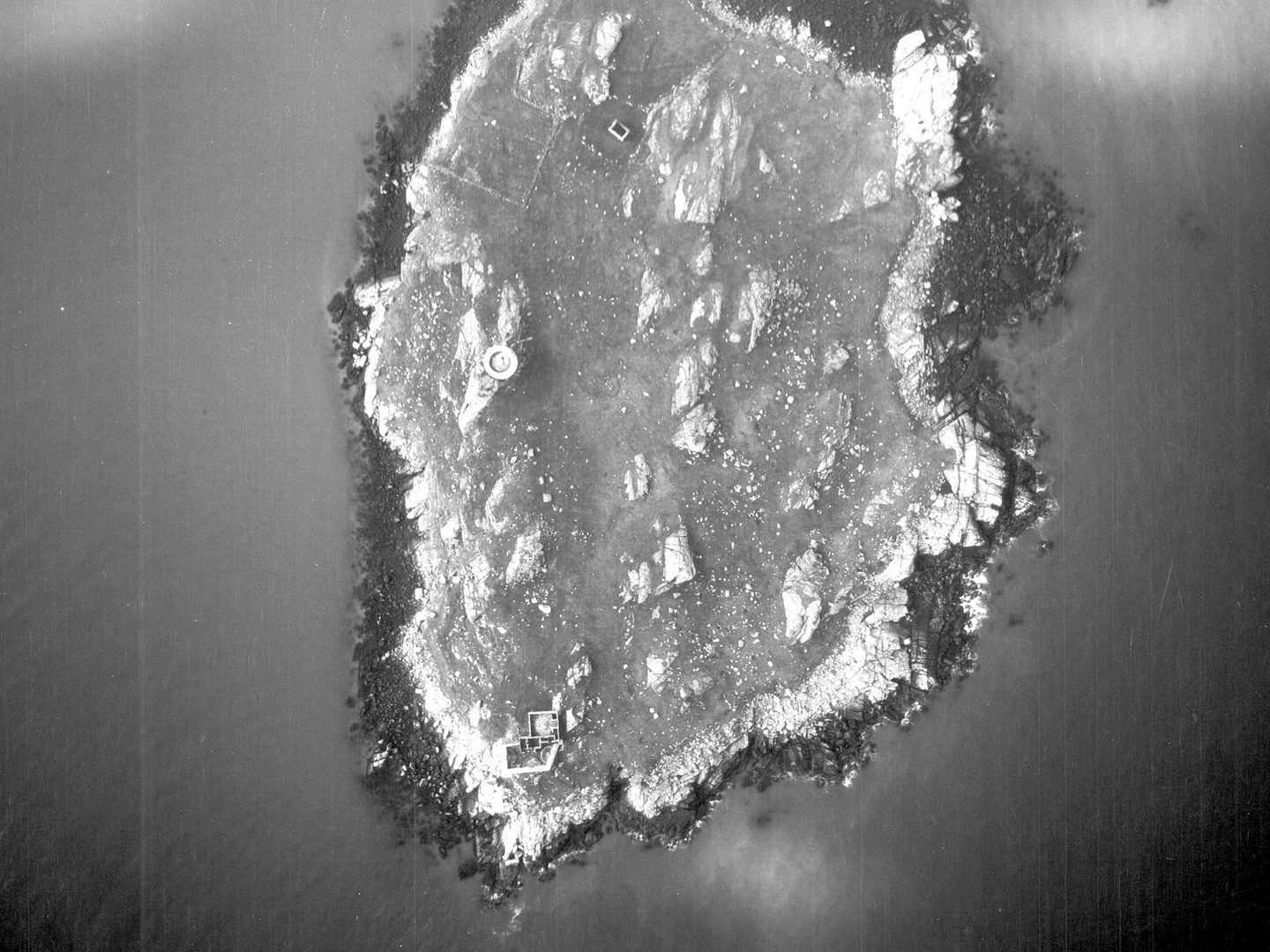 A historic black and white vertical aerial photo of a roughly oval-shaped island. A circular tower is visible to the left of centre, with a rectangular building complex at the bottom tip of the island.