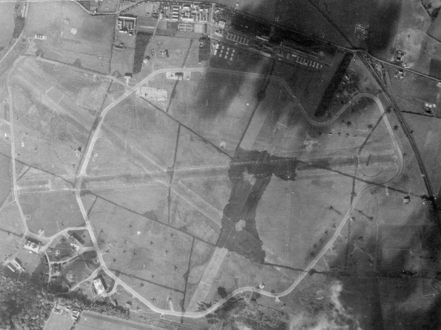A historic black and white vertical aerial photo of an airfield surrounded by fields. 3 camouflaged runways cross each other in the centre of the image. Airfield buildings are visible at the top of the image, with several aircraft hangars at the bottom.