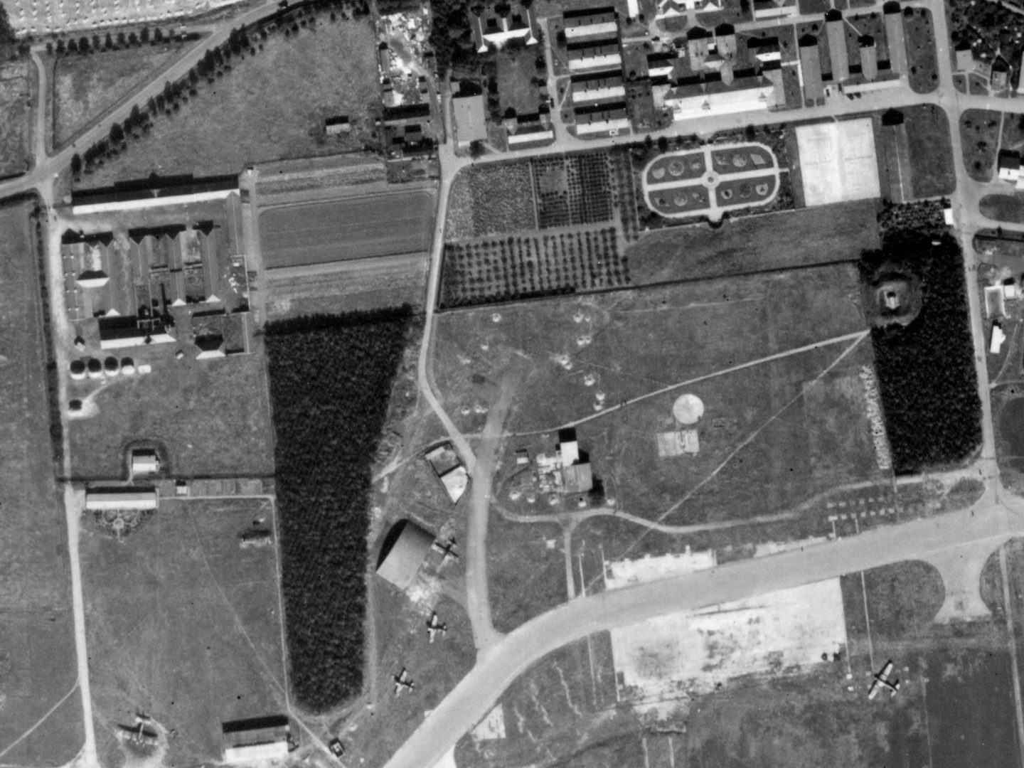 A historic black and white vertical aerial photo of a complex of buildings. A hangar and several aircraft are visible at the bottom of the image. Towards the top are several areas of planting, including a geometric formal garden.