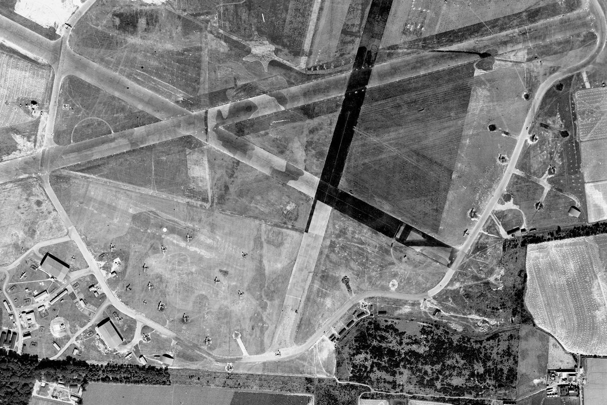 A historic black and white vertical aerial photo of an airfield surrounded by fields. 3 camouflaged runways cross each other in the centre of the image. Several aircraft hangars at the bottom left, along with several aircraft.