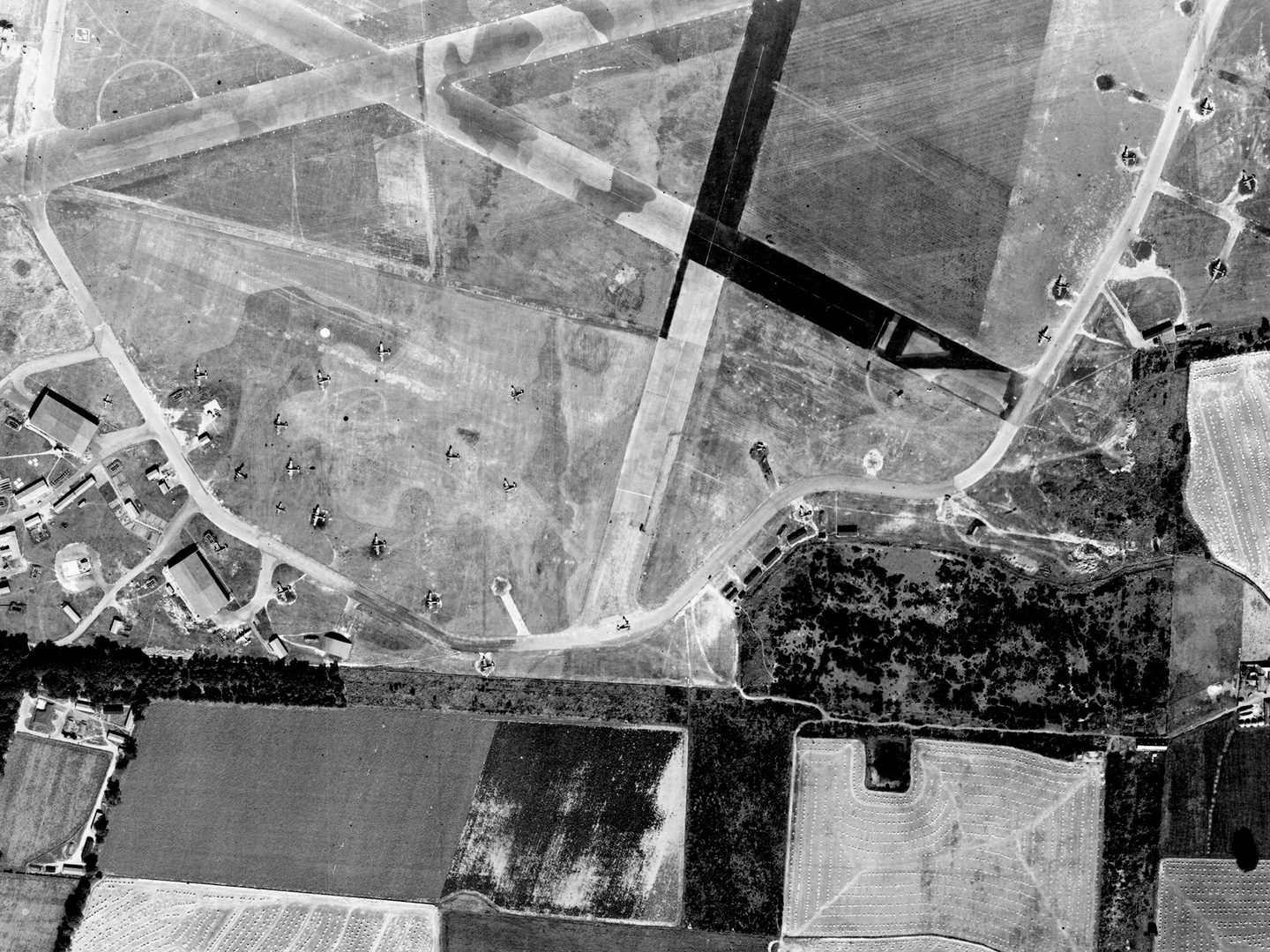 A historic black and white vertical aerial photo of an airfield surrounded by fields. 3 camouflaged runways cross each other at the top centre of the image. Several aircraft hangars are visible at the left-hand edge of the image, with numerous aircraft also visible.