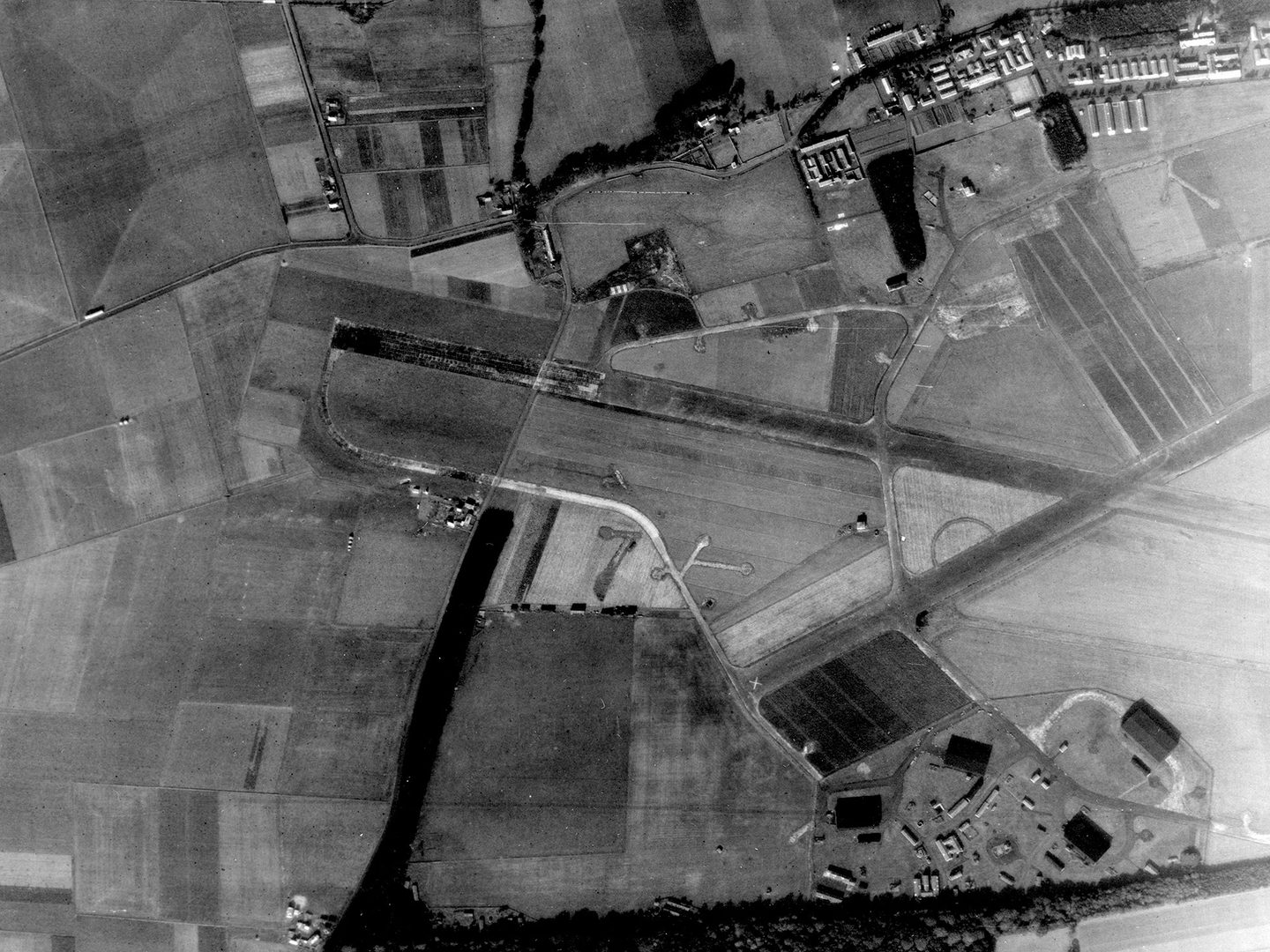 A historic black and white vertical aerial photo of an airfield surrounded by fields. 2 runways cross each other towards the right of the image. Airfield buildings are visible in the top right corner.