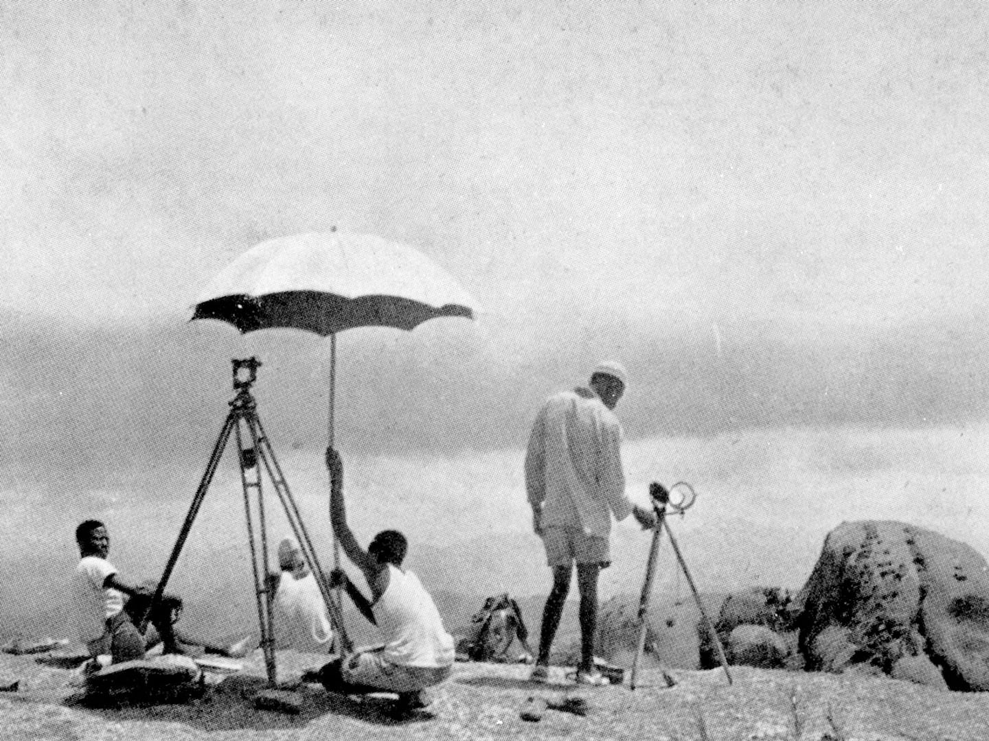 A black and white historic photo of 4 men standing on a mountaintop using surveying equipment. 1 man holds up a pale umbrella.
