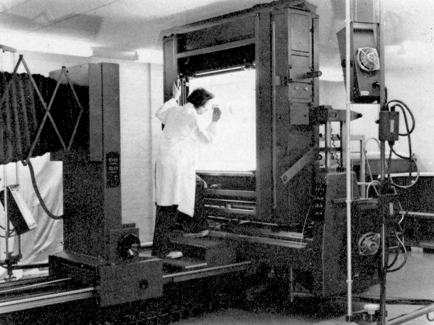 A black and white historic photo of a woman in a white lab coat operating a room-sized camera.