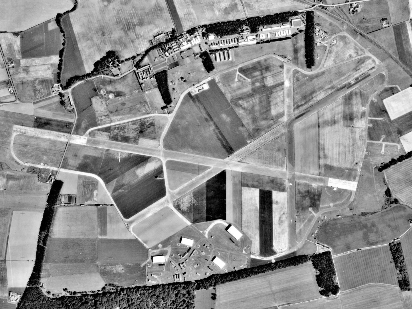 A historic black and white vertical aerial photo of an airfield surrounded by fields. 3 runways cross each other in the centre of the image. Airfield buildings are visible at the top of the image, with several aircraft hangars at the bottom.