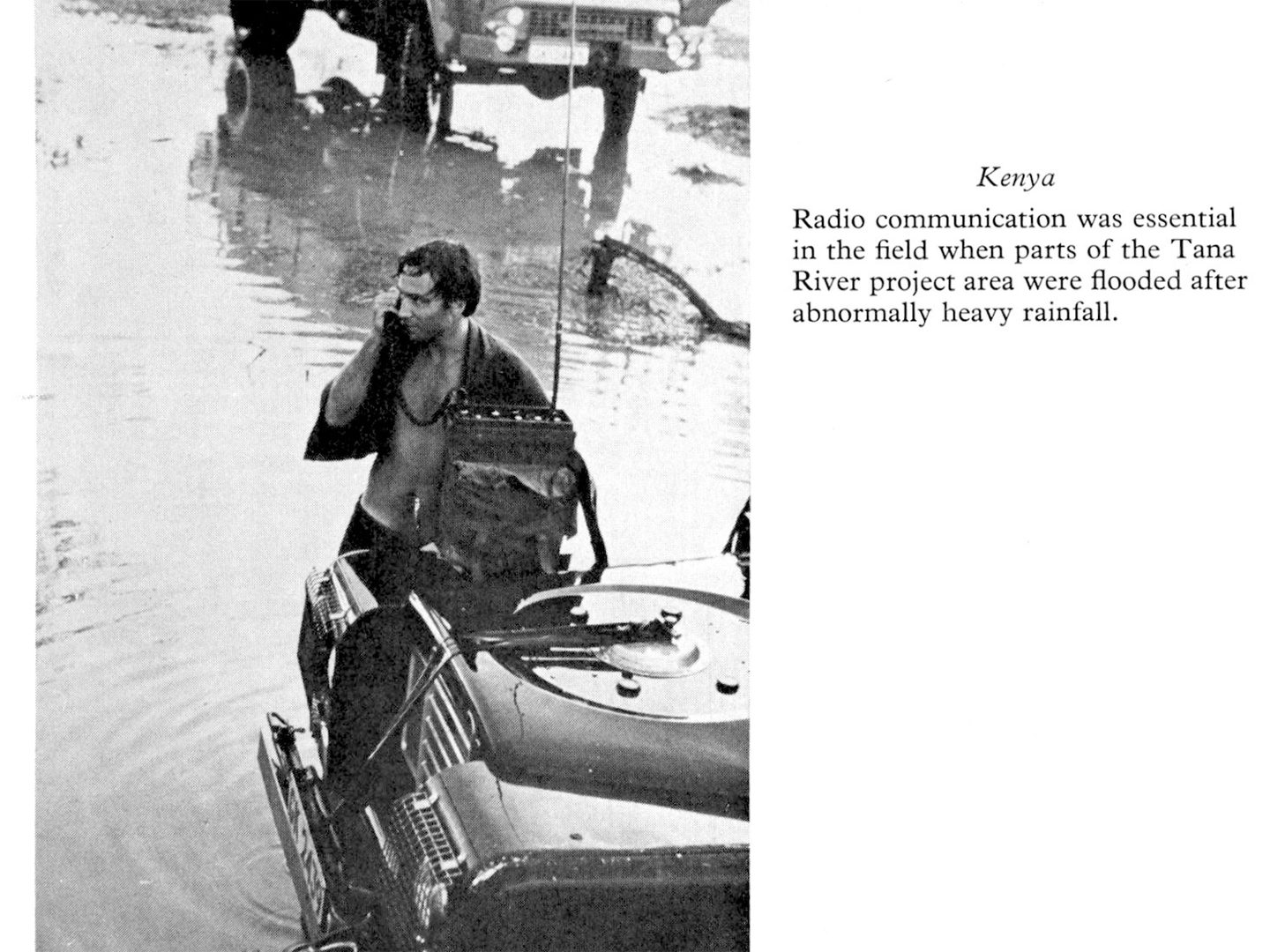 A black and white historic photo of a man in an open shirt, standing in water, using a telephone that is resting on the bonnet of a jeep.