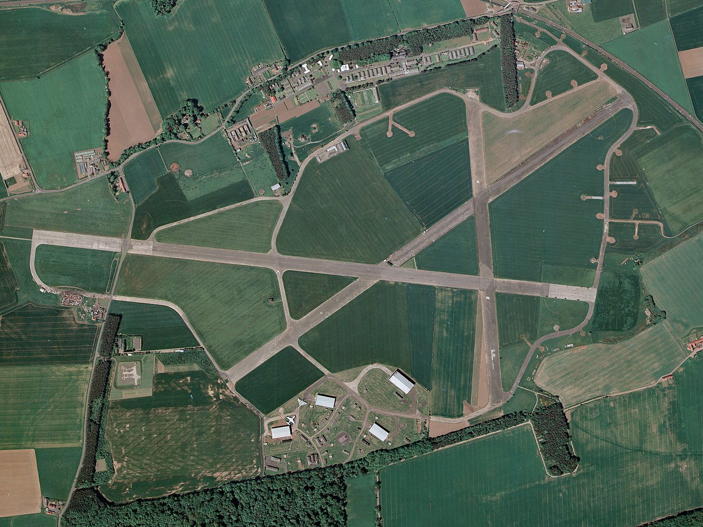 A historic colour vertical aerial photo of an airfield surrounded by fields. 3 runways cross each other in the centre of the image. Airfield buildings are visible at the top of the image, with several aircraft hangars at the bottom.